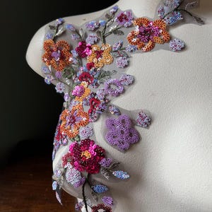 以下が含まれることがあります： オレンジ、ピンク、紫の色合いのカラフルなスパンコールが付いた花のアップリケ。花は透明なグレーのメッシュの裏地に付いています。デザインはマネキンに表示され、刺繍の複雑なディテールを示しています。