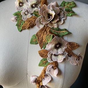 May include: An embroidered floral applique featuring detailed flowers in shades of brown, pink, and cream, accented with green and brown leaves. The applique is designed to be sewn onto fabric for decorative purposes.
