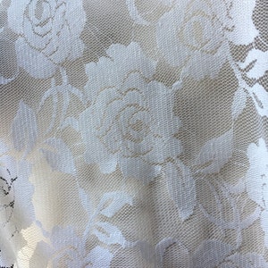 May include: Close-up of white lace fabric featuring a floral rose pattern. The delicate lace has a fine mesh background, with the rose designs standing out in a slightly raised texture. The fabric is likely used for apparel or home decor.