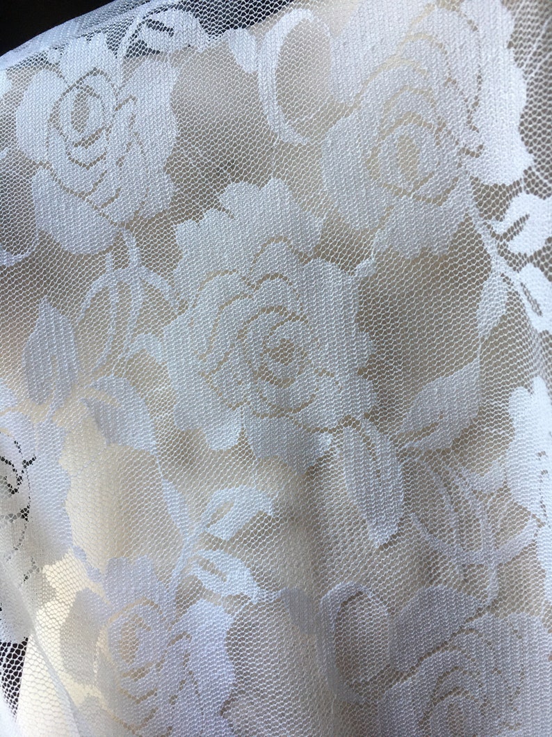 May include: Close-up of white lace fabric featuring a floral rose pattern. The delicate lace has a fine mesh background, with the rose designs standing out in a slightly raised texture. The fabric is likely used for apparel or home decor.
