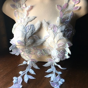 Może przedstawiać: Dwa kawałki białej i lawendowej aplikacji z koronki kwiatowej z opalizującymi cekinami. Koronka jest delikatna i ma wzór liści.