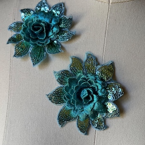 May include: Two teal sequined flower appliques. Each applique features a layered, embroidered flower with a central bloom and surrounding petals. The petals are edged with sequins, creating a shimmering effect. These appliques are ideal for embellishing garments or accessories.
