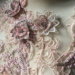 May include: Close-up of a delicate lace fabric with pink and beige floral appliques. The fabric is adorned with small beads and sequins, creating a luxurious and intricate design.