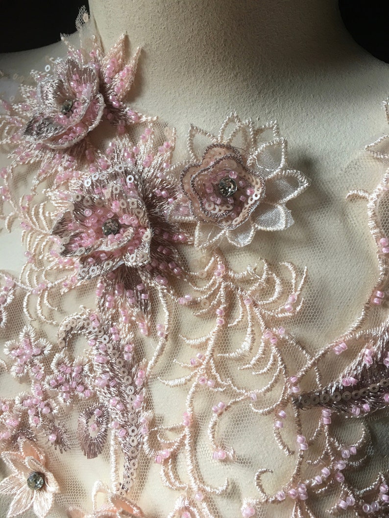 May include: Close-up of a delicate lace fabric with pink and beige floral appliques. The fabric is adorned with small beads and sequins, creating a luxurious and intricate design.