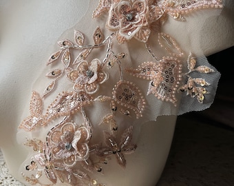 May include: A delicate floral applique with a blush pink color and gold sequins. The applique features a cluster of flowers and leaves, perfect for adding a touch of elegance to a garment or craft project.