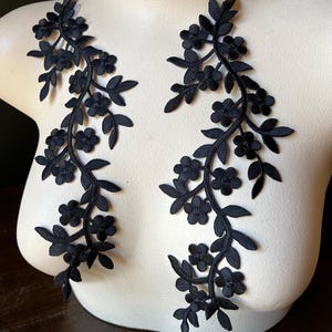 Puede incluir: Dos apliques de encaje floral negro. Cada aplique presenta un delicado diseño de vid con pequeñas flores y hojas. Los apliques se exhiben en un maniquí blanco, mostrando sus intrincados detalles y su potencial para el embellecimiento de prendas.