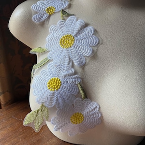 May include: A white sheer fabric with four embroidered white daisies with yellow centers. The daisies are arranged in a vertical line with green stems and leaves.
