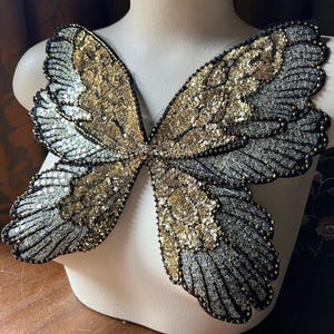 Puede incluir: Un par de alas de mariposa de lentejuelas doradas y plateadas. Las alas están unidas a un busto de maniquí blanco.