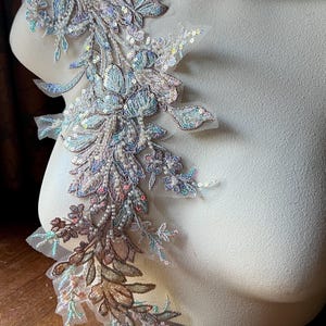 May include: An ornate floral applique with iridescent sequins and beads. The design features leaves and flowers in shades of blue, silver, and brown, attached to a sheer mesh backing. The applique is displayed on a mannequin.
