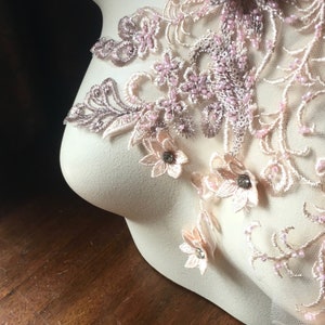 May include: A delicate lace applique with pink and beige floral designs and small pearl-like beads. The applique is displayed on a white mannequin.