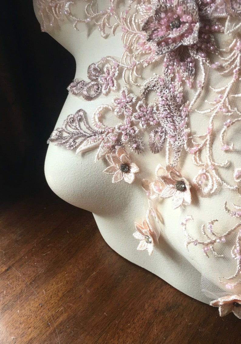 May include: A delicate lace applique with pink and beige floral designs and small pearl-like beads. The applique is displayed on a white mannequin.
