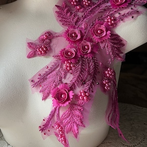 Könnte beinhalten: Fuchsiafarbene Blumenapplikation mit aufwendigen Details. Die Applikation hat Stoffblätter, kleine Blüten und Kunstperlen. Das Design ist auf einem transparenten Netzrücken befestigt. Geeignet für verschiedene Bastelprojekte.