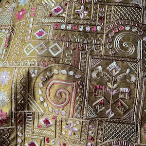 May include: Close-up of a section of fabric with detailed embroidery. The base fabric is a muted gold colour, and the embroidery features geometric and floral patterns in gold, pink, and white. The stitching is intricate, creating a textured surface.