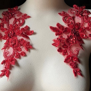 May include: Two red lace appliques with beaded embellishments. The appliques are on a white background and are perfect for adding a touch of elegance to any garment.
