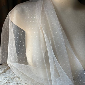 May include: White dotted tulle fabric draped over a dress form. The fabric is sheer and has a delicate, feminine look.