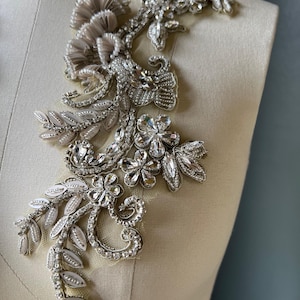 May include: A detailed silver applique with floral and leaf patterns, embellished with sparkling crystals and beads. The decorative piece is displayed on a mannequin, highlighting its potential use in fashion. The design features intricate detailing and a luxurious texture.