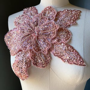 May include: A large, pink floral applique with intricate sequin and bead detailing. The applique is displayed on a mannequin, showcasing its shape and design. The petals are a soft pink, with a lighter center.