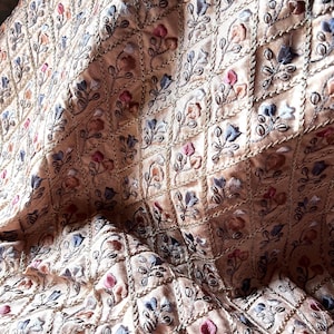 May include: Close-up of a beige fabric with an embroidered floral pattern. The embroidery features small flowers in shades of pink, blue, and orange, set within a grid of stitched squares. The fabric has a subtle sheen, suggesting a luxurious material.