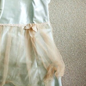 May include: A light blue dress with a layered design. The dress features a sheer, peach-colored overlay and a delicate floral embellishment at the waist. The dress has a satin-like texture and a lace trim.