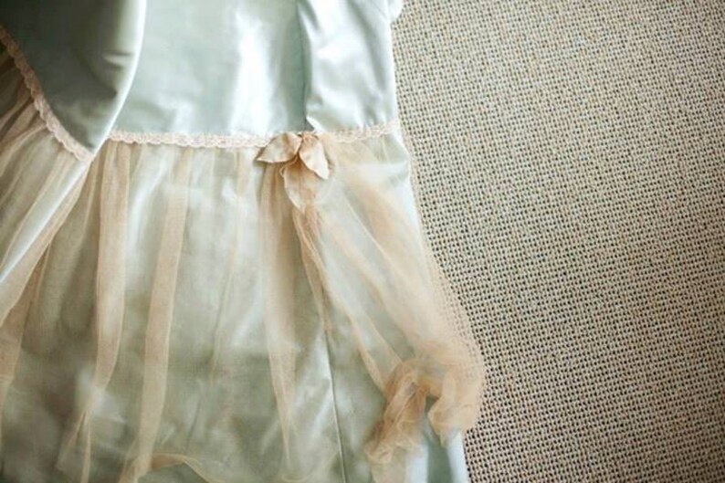 May include: A light blue dress with a layered design. The dress features a sheer, peach-colored overlay and a delicate floral embellishment at the waist. The dress has a satin-like texture and a lace trim.