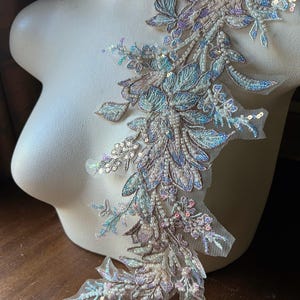 May include: An intricate, beaded floral applique in shades of blue, lavender, and cream. The delicate design features leaves, flowers, and sequins, creating a shimmering, textured effect. The applique is displayed on a mannequin, showcasing its potential for embellishment.