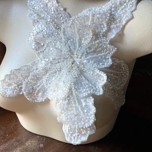 Pode incluir: Aplique floral branco com detalhes intrincados de lantejoulas. O aplique é exibido em um manequim, mostrando seu design e textura. As lantejoulas capturam a luz, criando um efeito brilhante. Esta peça decorativa é adequada para adornar roupas ou acessórios.