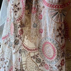 Könnte beinhalten: Nahaufnahme eines transparenten Stoffes mit aufwendiger Stickerei. Das Design zeigt florale und geometrische Muster in rosa, blauen und goldenen Fäden. Der Stoff ist cremefarben, und die Stickerei ist mit Pailletten und Perlen verziert.