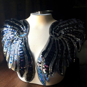 May include: A pair of black and blue sequined wings on a white mannequin. The wings are made of a sheer fabric with sequins sewn on in a feather pattern.