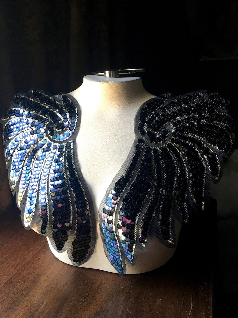 May include: A pair of black and blue sequined wings on a white mannequin. The wings are made of a sheer fabric with sequins sewn on in a feather pattern.