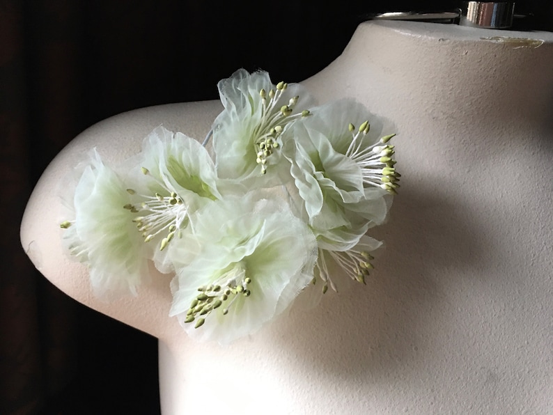 May include: A cluster of delicate, white, sheer fabric flowers with green centers. The flowers are arranged in a circular shape and are attached to a white surface.