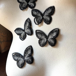 May include: Five black butterfly patches with silver embroidered details are arranged on a white surface. The butterflies have a delicate, detailed design, with their wings spread. These patches could be used for clothing or craft projects.