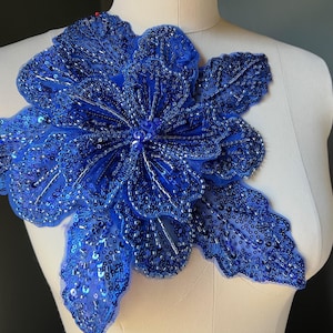 May include: A large, royal blue floral applique with intricate bead and sequin detailing. The applique is shaped like a flower with multiple petals and leaves, perfect for embellishing garments or accessories.