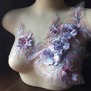 Op de afbeelding: Een delicate, bloemen applicatie in tinten lavendel, roze en lichtblauw. Het ontwerp toont geborduurde bloemen met kralen in het midden en veervormige accenten, alles op een transparante mesh achtergrond. Dit decoratieve stuk is waarschijnlijk bedoeld om kleding of accessoires te verfraaien.