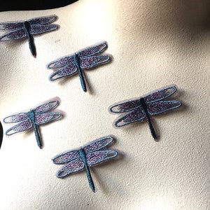 May include: Five embroidered dragonfly patches. Each patch features a dragonfly with purple and blue wings, a black body, and a blue tail. The patches are arranged on a light-colored surface.