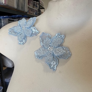 Może przedstawiać: Dwa jasnoniebieskie aplikacje kwiatowe z koralików z kryształowym środkiem. Aplikacje znajdują się na białym tle.