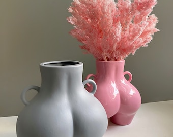 May include: Two ceramic vases shaped like human torsos with handles on the sides. One vase has a matte gray finish, while the other is glossy pink and contains a bouquet of fluffy, light pink dried foliage extending from the top opening.