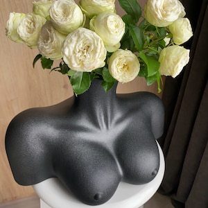 May include: A black bust-shaped vase filled with white roses. The vase is a matte black and has a detailed form. The roses are a creamy white color with green leaves. The vase is sitting on a white stool.