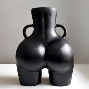 Peut inclure: Un vase en céramique noir avec une forme unique, rappelant une silhouette humaine vue de dos. Le vase a une finition mate et lisse, deux petites anses et une base large. Le design est moderne et sculptural.