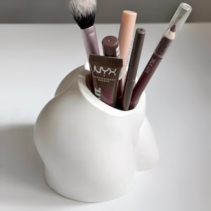 Op de afbeelding: Een witte keramische make-up borstelhouder in een unieke vorm, gevuld met diverse cosmetica-artikelen. De houder bevat een make-up borstel, lippenpotloden en make-up tubes. De tekst "NYX PROFESSIONAL MAKEUP" is zichtbaar op een van de tubes.