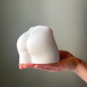 May include: A white ceramic sculpture of a human form's lower back and buttocks. The sculpture is held in a hand, showcasing its smooth, curved form. The piece is a minimalist art object, likely for home decor.