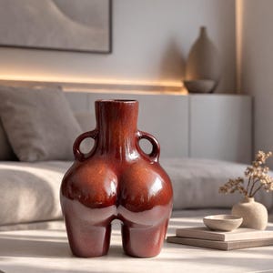 May include: A glossy, deep reddish-brown ceramic vase with a rounded, dual-lobed lower body and two small, circular handles attached to the neck. The surface has a reflective, smooth finish that catches the light, highlighting its curved form.