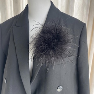 May include: A black blazer with a large, fluffy black feather accent pinned to the lapel. The blazer has a classic double-breasted design with two buttons and a notched collar. The background is a neutral color.