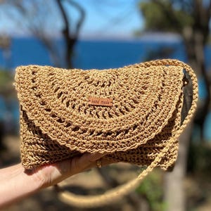 May include: A tan, hand-crocheted clutch bag with a flap closure. The bag features an open weave design and a small, rectangular tag that reads "hand made". The strap is a thin, braided cord.