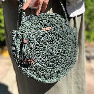 May include: A green crocheted round bag with a brown leather tag that says "hand made" and brown wooden beads on a tassel.