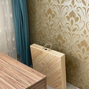 May include: A wooden box with a diagonal pattern on the lid. The box is closed and has a rope handle. The box is sitting on a light wood floor in front of a wall with a gold and white damask pattern.