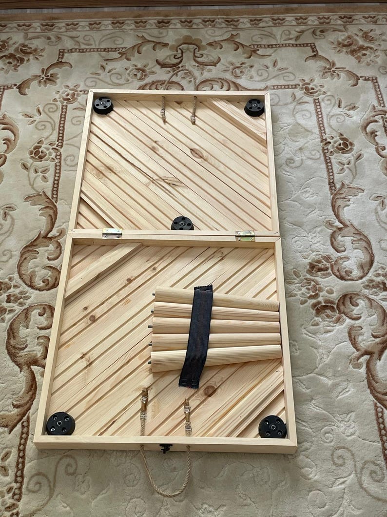 May include: A wooden game board with a rope handle and black rubber feet. The board is open and shows a set of wooden dowels with a black strap.