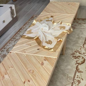 May include: A wooden table with a chevron pattern top. The table is made of light wood and has a natural finish. A white sheer scarf with gold trim and floral embroidery is draped over the table.