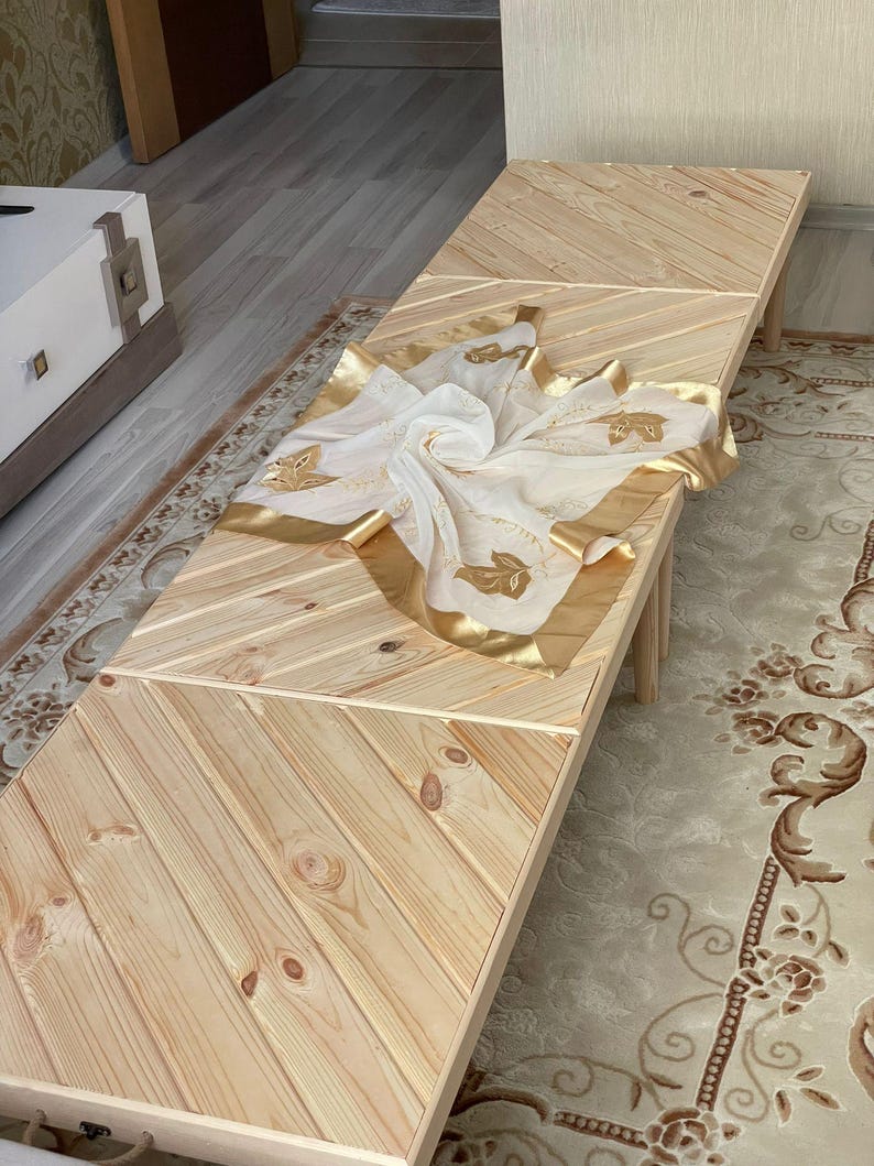 May include: A wooden table with a chevron pattern top. The table is made of light wood and has a natural finish. A white sheer scarf with gold trim and floral embroidery is draped over the table.