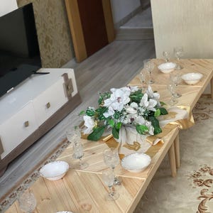 May include: A wooden table set with white bowls and clear glass cups. The table is made of light wood planks and has a chevron pattern. A bouquet of white flowers is in the center of the table.