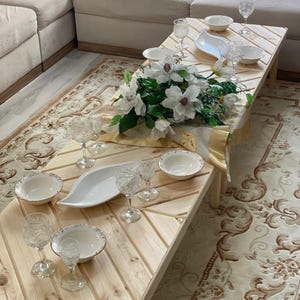 May include: A wooden table with a chevron pattern, set for a meal with white bowls, plates, and glasses. A bouquet of white flowers sits in the center of the table.